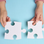 Female hand holds two large paper empty white jigsaw puzzles on a blue background, top view