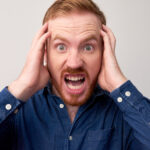 Portrait of red haired young man in panic holding his head and looking at camera